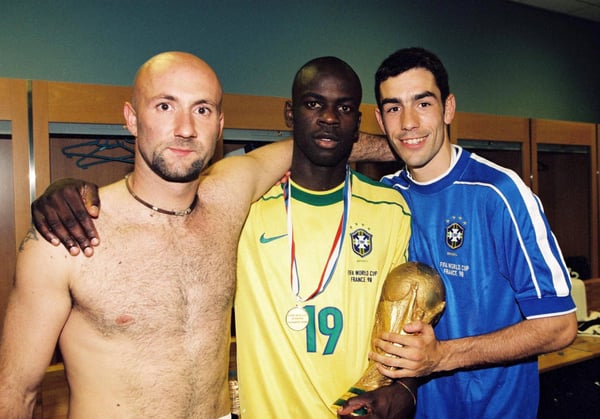 世界杯FIFA体育新闻：利利安·图拉姆身穿巴西球衣，手捧奖杯，在法国队赢得1998年国际足联世界杯决赛后，与法比安·巴特兹和罗伯特·皮雷斯合影。